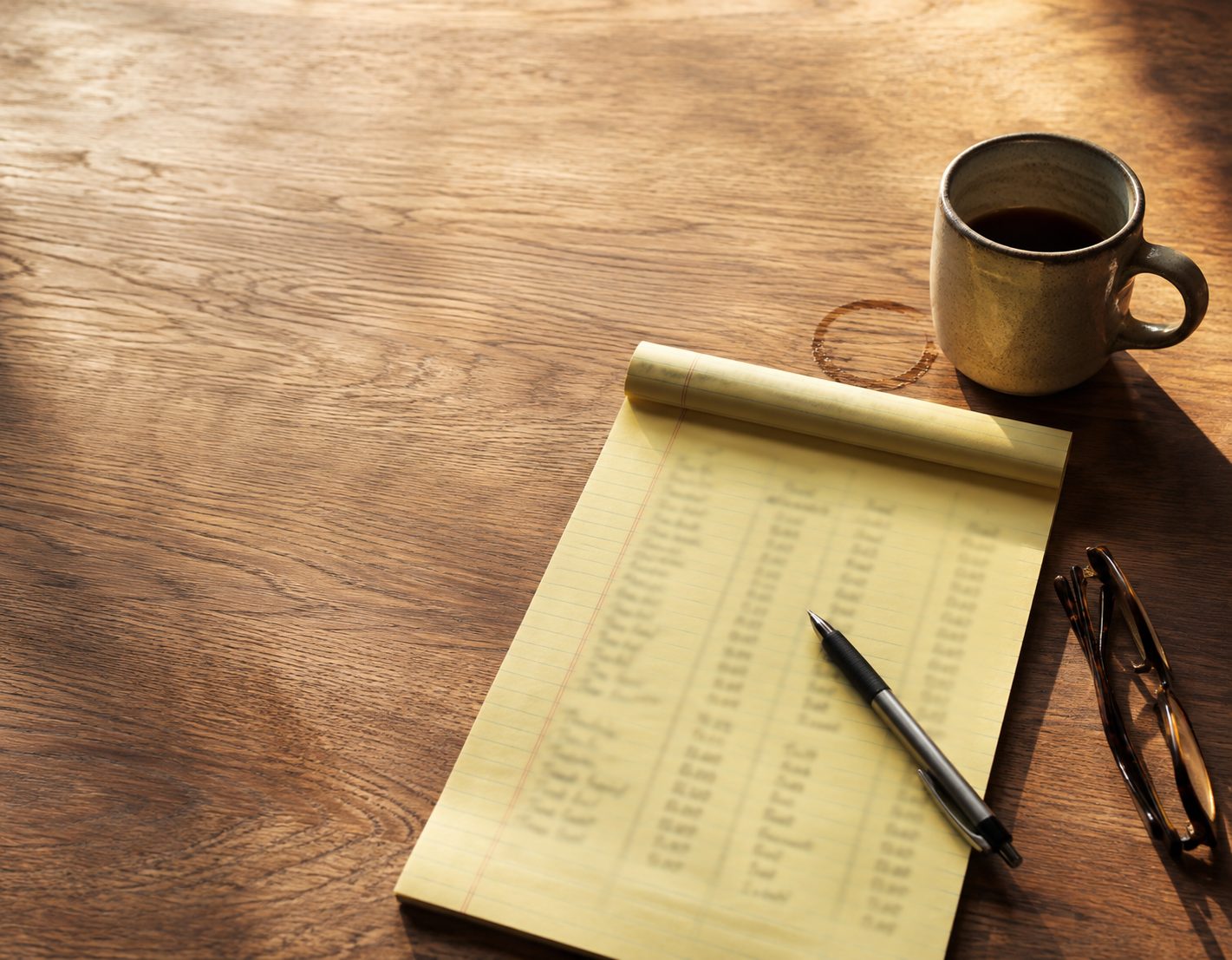 A yellow legal pad on a wooden table with columns of handwritten figures, a pen, reading glasses and a mug of coffee