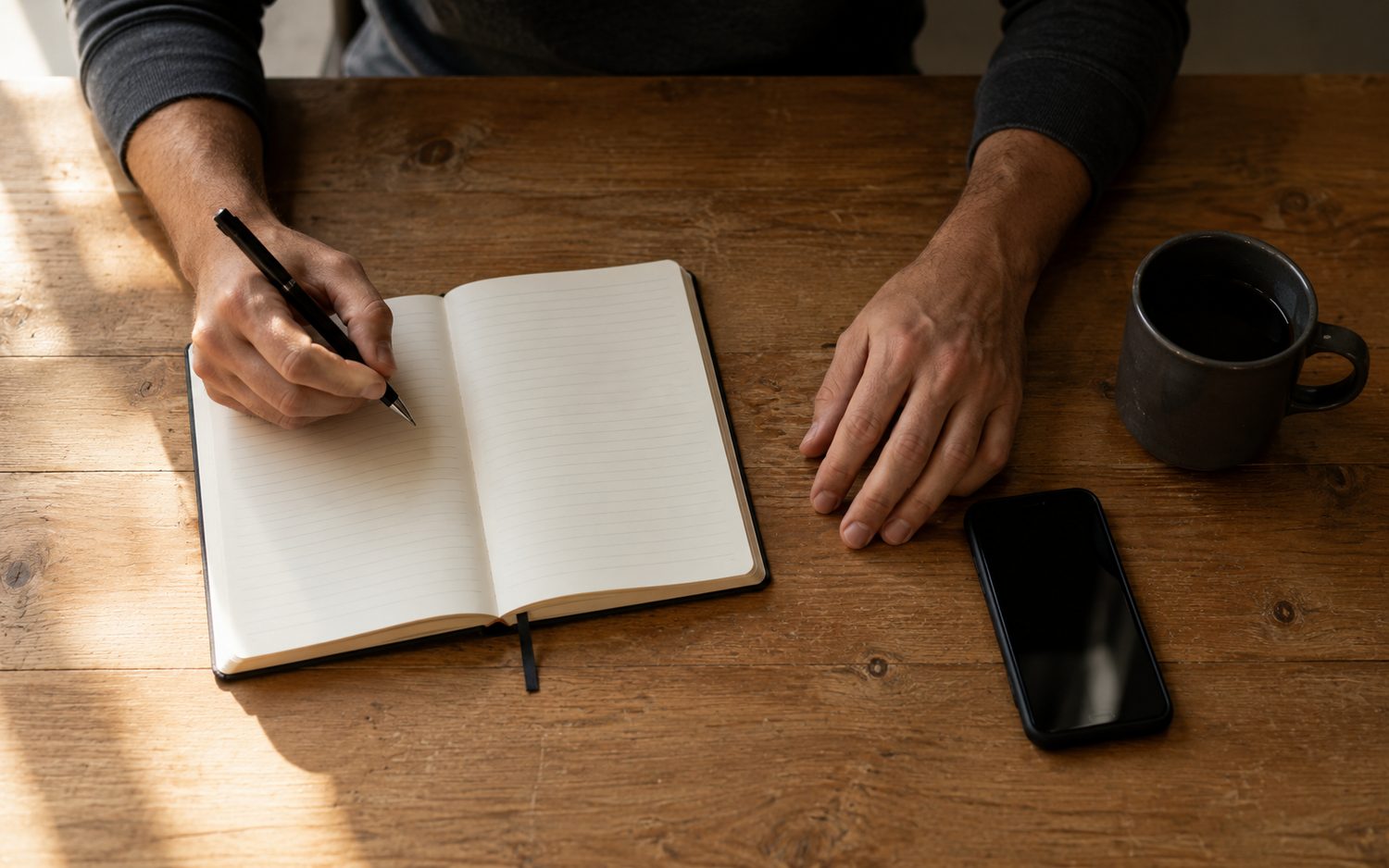 A person's hands at a wooden table, writing in a notebook beside a phone and a mug